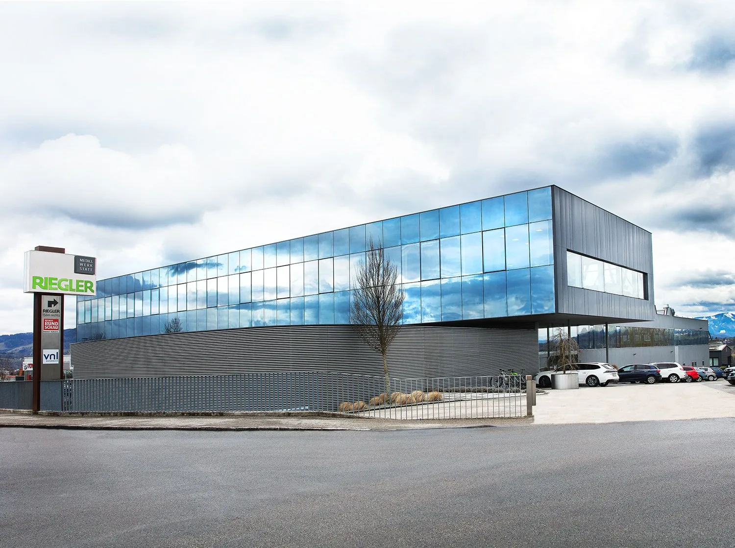 Bürogebäude mit spiegelnder Glasfassade und RIEGLER-Schild an der Straße, blauer Himmel
