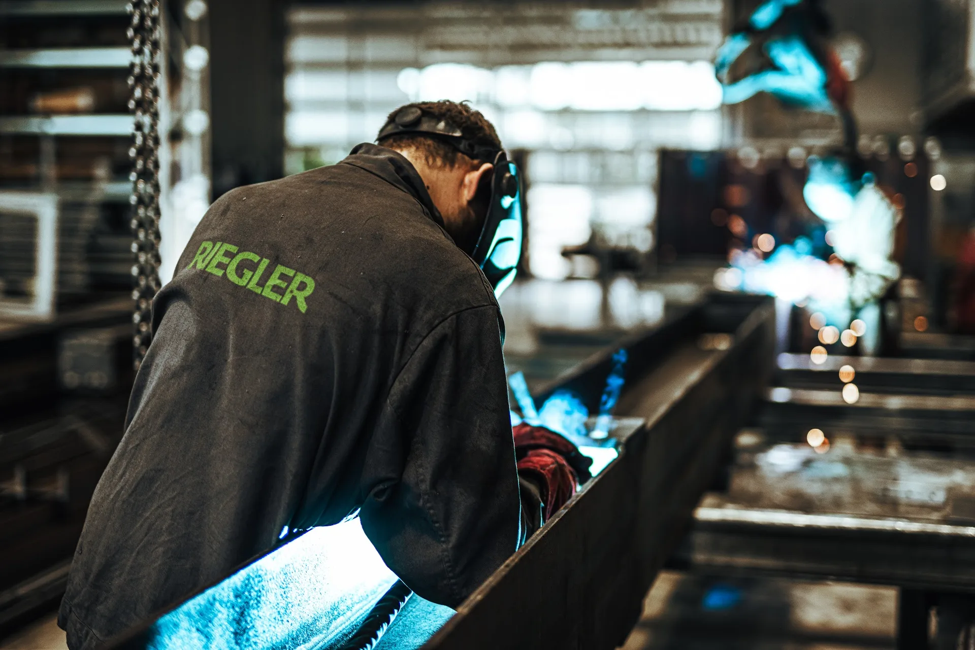 Mitarbeiter in schwarzer Jacke mit grünem RIEGLER-Schriftzug beim Schweißen in der Werkhalle, blaues Licht