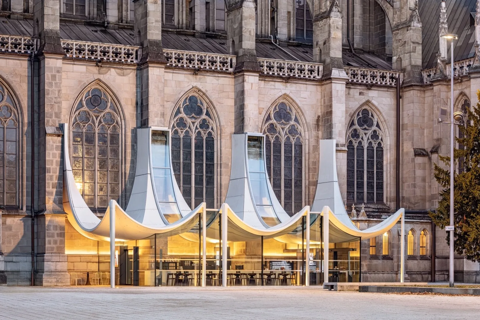 Gläserner Pavillon mit geschwungenen weißen Dachelementen vor der gotischen Fassade des Mariendoms in Linz, abends beleuchtet