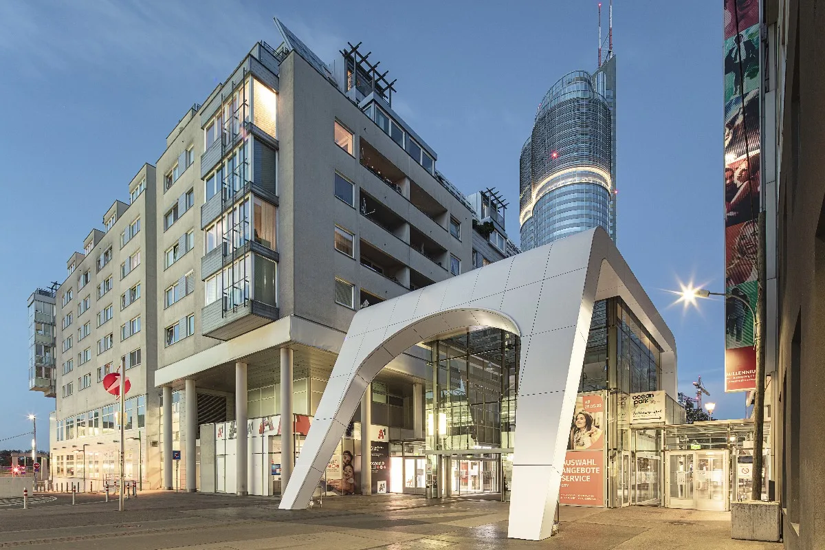 Weiß verkleideter Eingangsbogen an der Straßenseite in der Dämmerung, dahinter ein Hochhaus